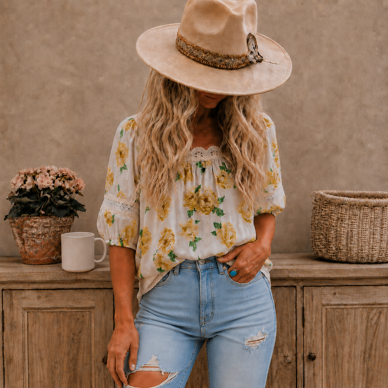A woman wearing a cream floral bohemian blouse with yellow flowers and green leaves, paired with light-wash distressed jeans and a beige wide-brim hat, standing in front of a rustic wooden wall.