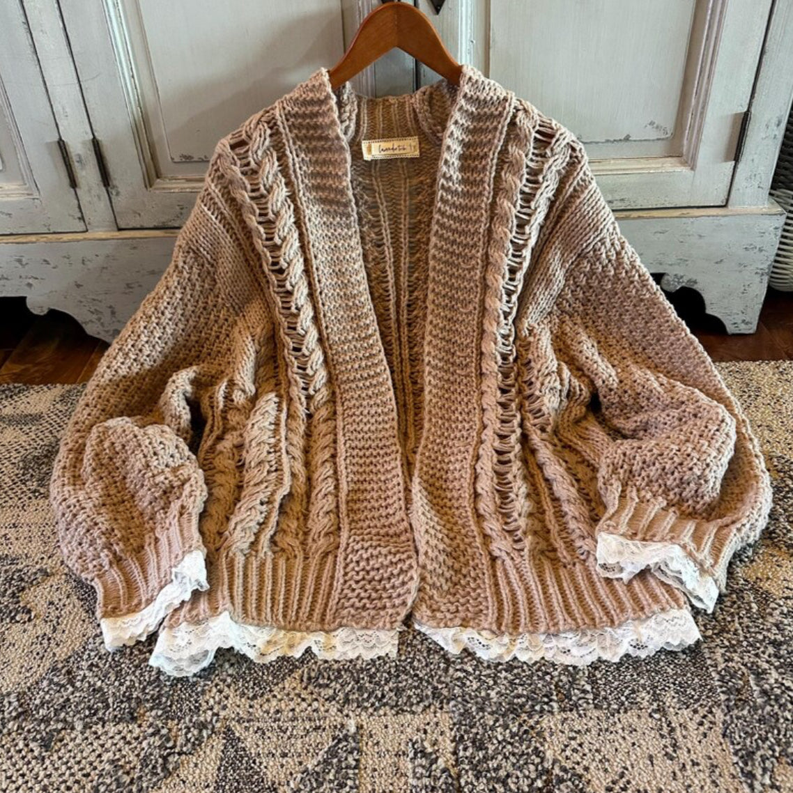 Knitted cardigan with lace trim on a wooden hanger against a rustic wooden cabinet background.