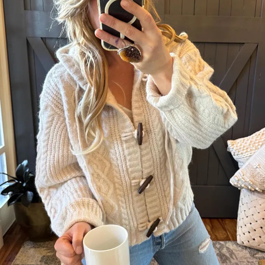 Person wearing a beige cardigan with buttons, holding a phone and a mug indoors.