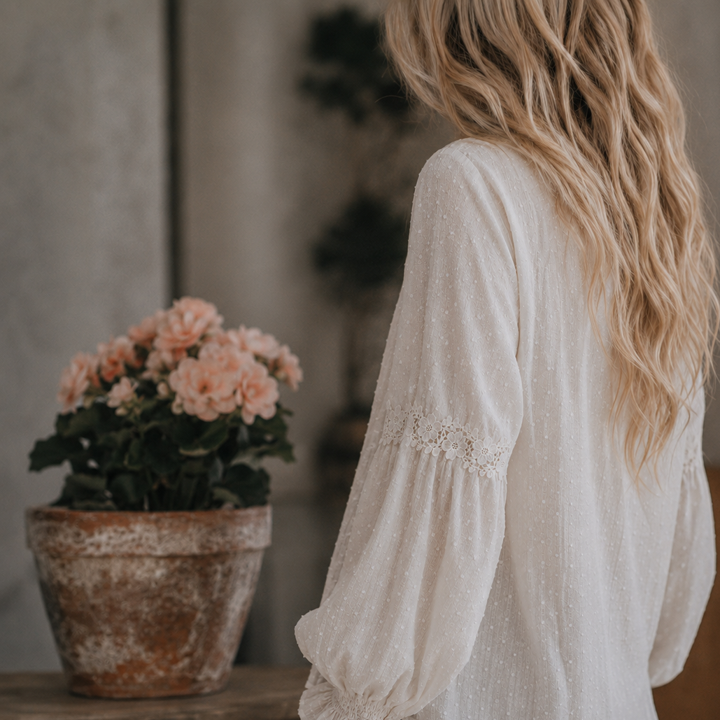 A woman with long blonde hair is shown from behind wearing a white long-sleeve blouse with lace detailing on the upper sleeve and gathered cuffs, paired with dark blue jeans. She is standing next to a rustic wooden table holding a potted pink flower in a textured pot.