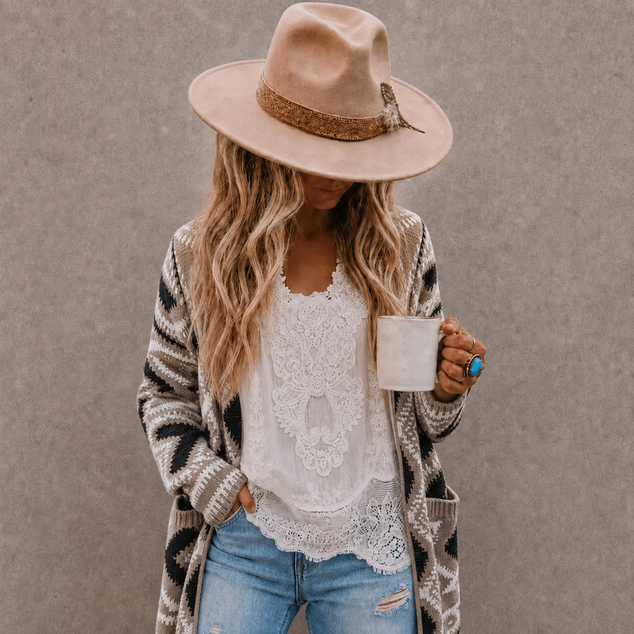A woman wearing a beige wide-brim fedora hat, a black and beige striped knit cardigan, a white lace top, and blue jeans, holding a white mug against a textured gray wall.