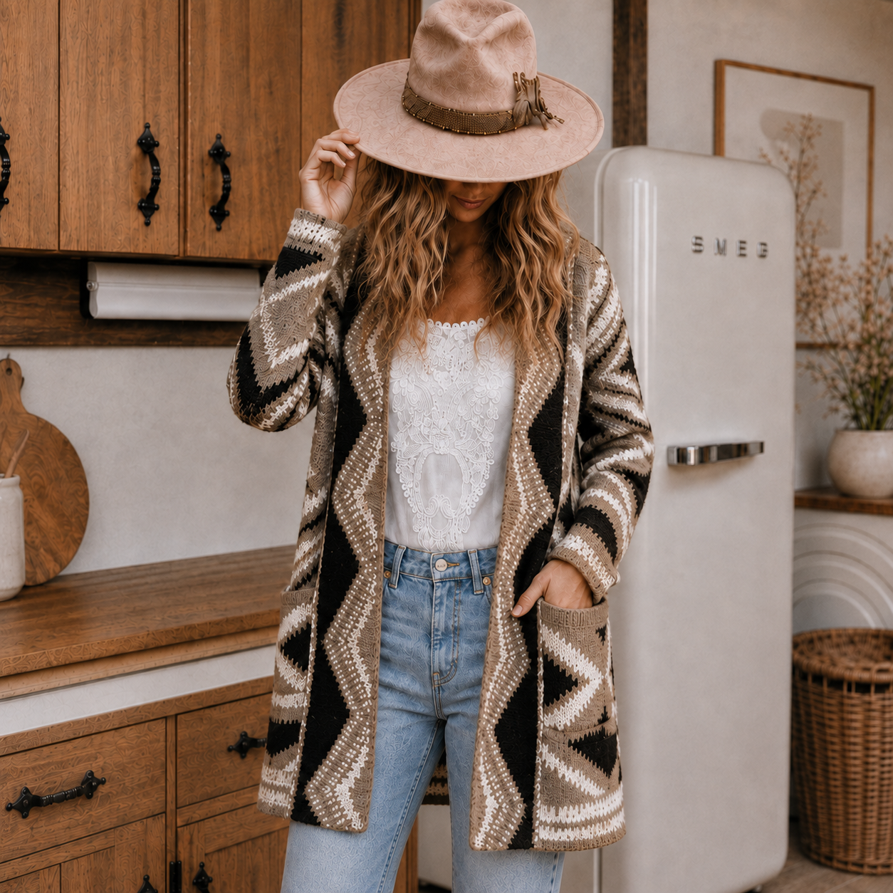 A woman stands in a rustic kitchen wearing a long open-front knit cardigan with a beige, brown, and black geometric pattern, a white lace top, light blue jeans, and a wide-brimmed beige hat. A white SMEG refrigerator is visible in the background.