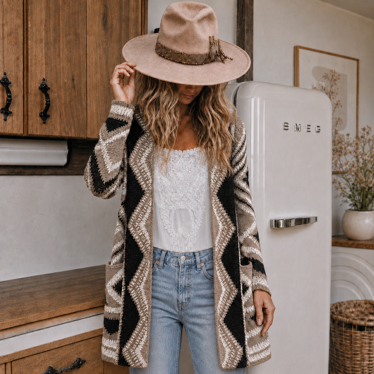 A woman stands in a kitchen wearing a long open-front knit cardigan with a beige, black, and white geometric zigzag pattern, a white lace top, light blue jeans, and a wide-brimmed beige hat.