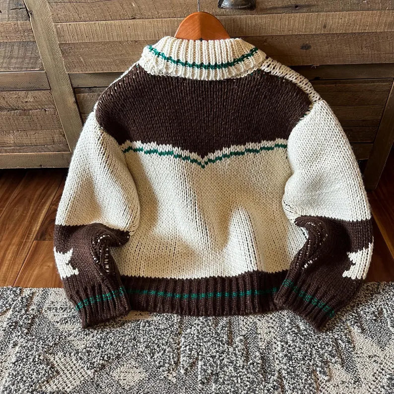 A hand-knit sweater with a brown and cream color-blocked design and thin green stripe accents, hanging on a wooden hanger against a rustic wooden wall.