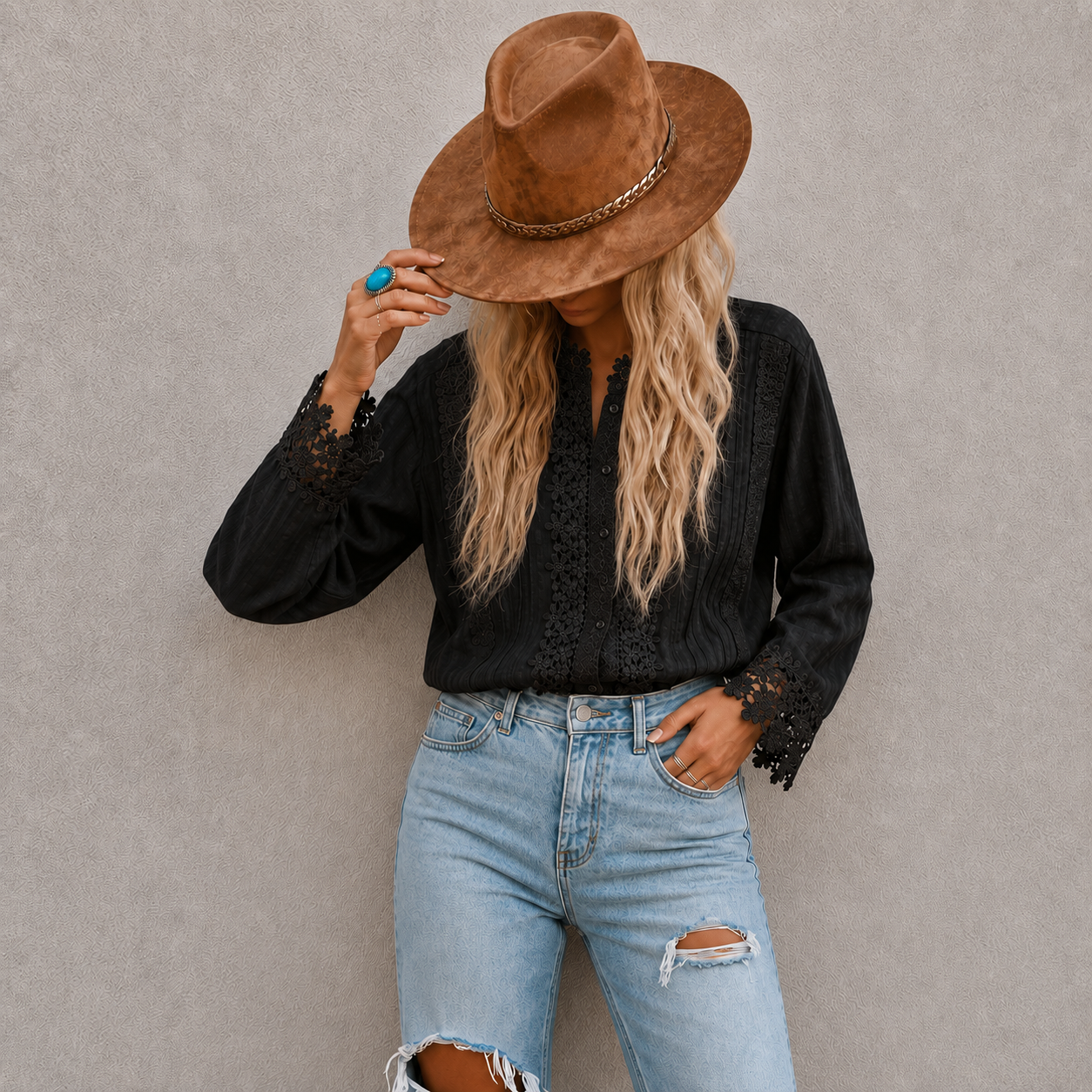 A woman wearing a brown wide-brim fedora hat, a black long-sleeve blouse with lace cuffs, and light blue ripped jeans, posing against a textured gray wall.