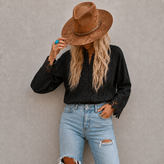 A woman wearing a brown wide-brim fedora hat, a black long-sleeve blouse with lace cuffs, and light blue ripped jeans, posing against a textured gray wall.