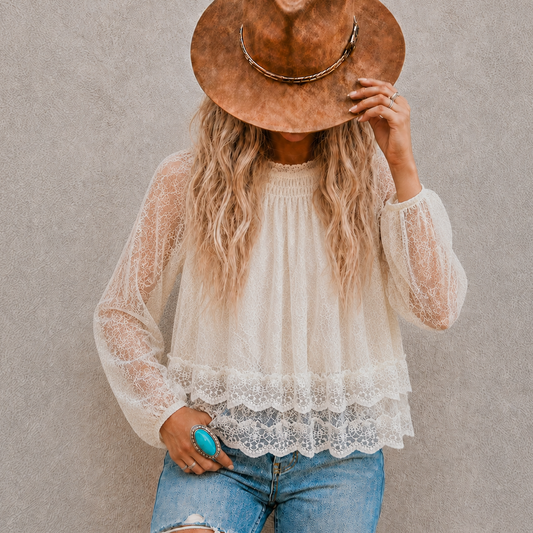 A woman wearing a cream lace long-sleeve blouse with a scalloped hem, blue jeans, and a brown wide-brim hat, standing against a textured gray wall.