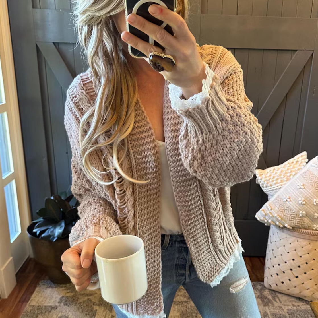 Person wearing a beige knitted cardigan holding a mug indoors.