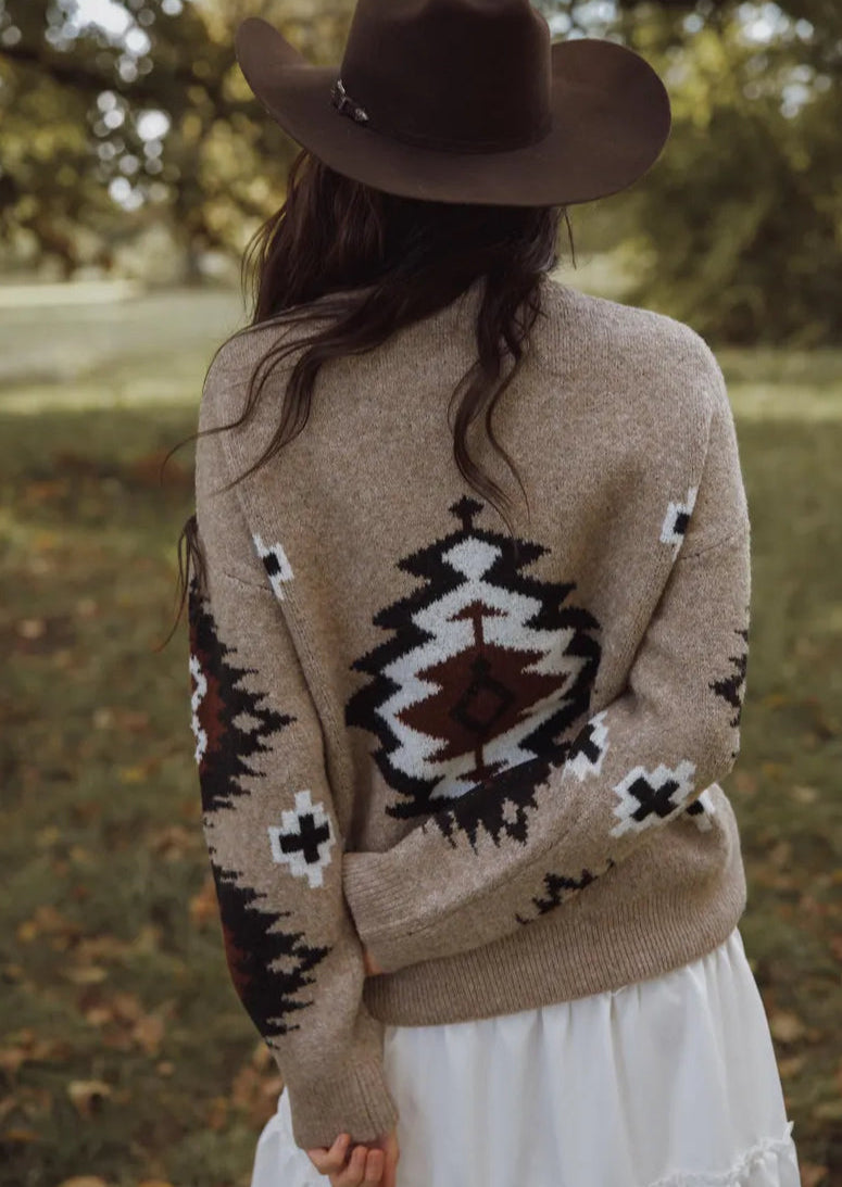 Person wearing a brown sweater with a geometric pattern and a cowboy hat, standing in a natural setting.