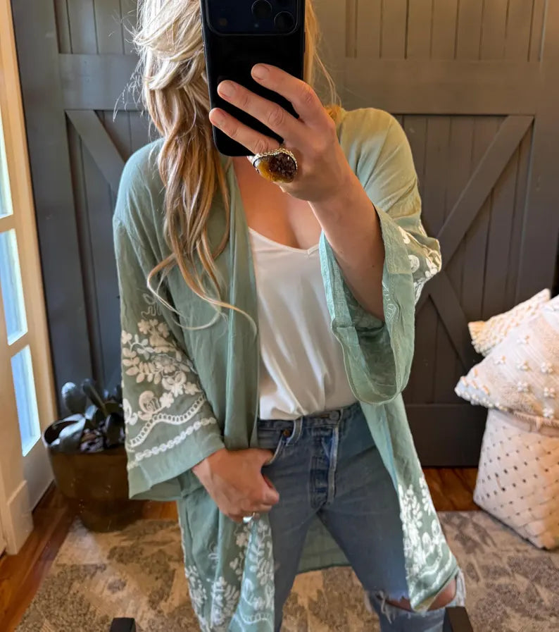 A woman takes a mirror selfie wearing a sage green open-front kimono cardigan with white floral lace embroidery on the sleeves and hem, layered over a white tank top and blue jeans.