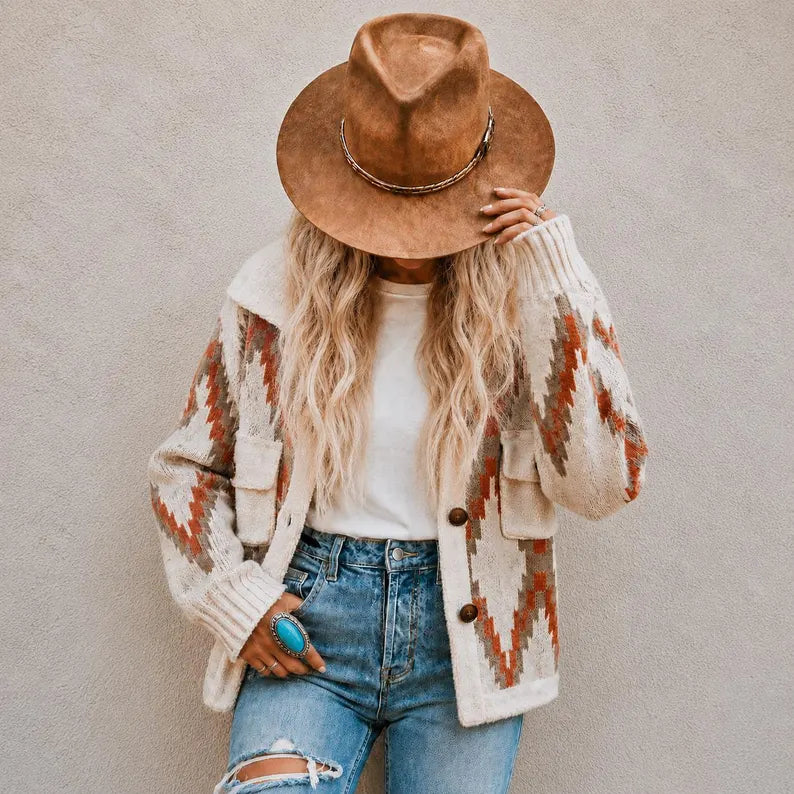 A woman wearing a brown wide-brim hat, a cream knit cardigan with orange and brown geometric patterns, a white t-shirt, and blue jeans, standing against a textured gray wall.