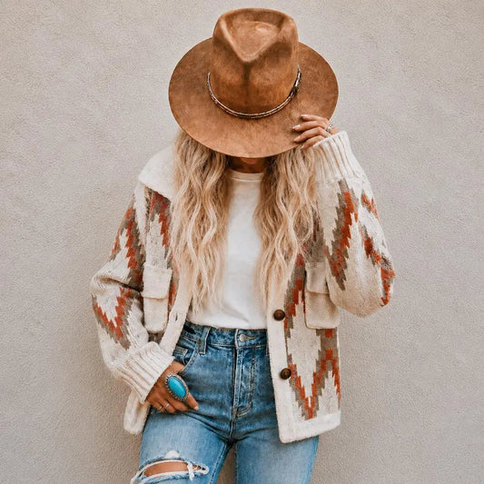 A woman wearing a brown wide-brim hat, a cream knit cardigan with orange and brown geometric patterns, a white t-shirt, and blue jeans, standing against a textured gray wall.