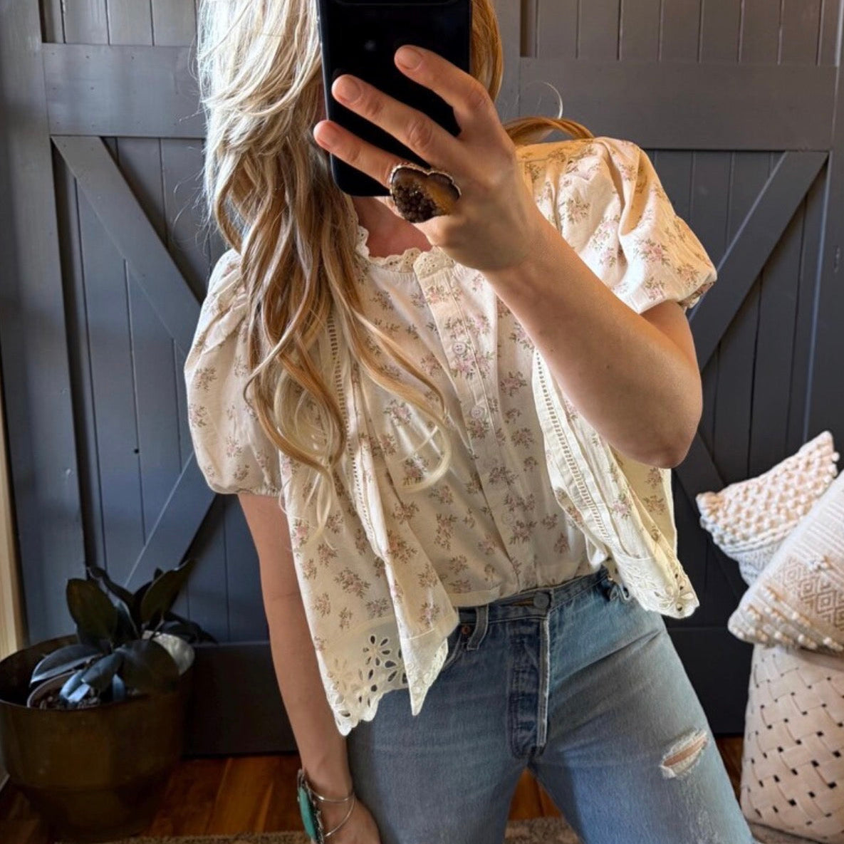 Person wearing a floral blouse and jeans, taking a mirror selfie.