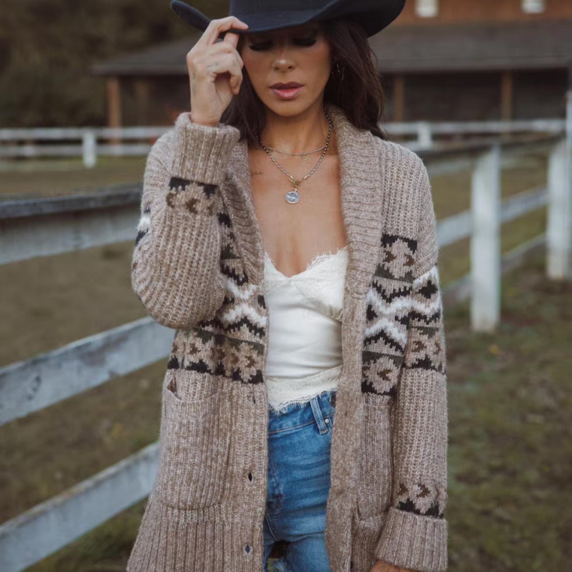Woman wearing a long beige cardigan with a pattern, white top, and blue jeans outdoors.