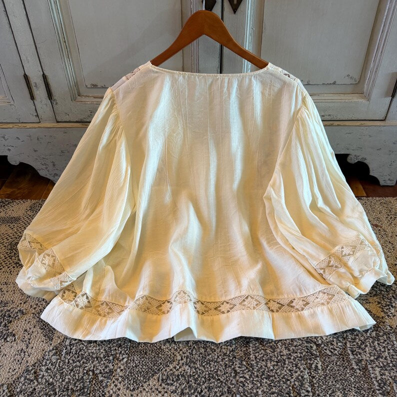 A cream-colored, long-sleeved blouse with lace trim on the cuffs and hem, hanging on a wooden hanger against a distressed white cabinet background.