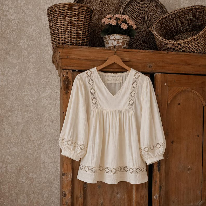 An off-white/cream bohemian-style blouse with embroidered geometric patterns, hanging on a wooden hanger from a rustic wooden cabinet with wicker baskets in the background.