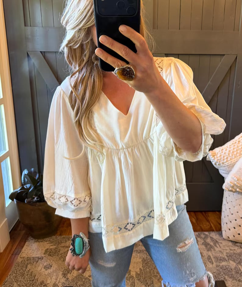 A woman takes a mirror selfie wearing a cream-colored bohemian-style blouse with puff sleeves, a V-neckline, and embroidered details on the cuffs and hem, paired with distressed light-wash jeans.