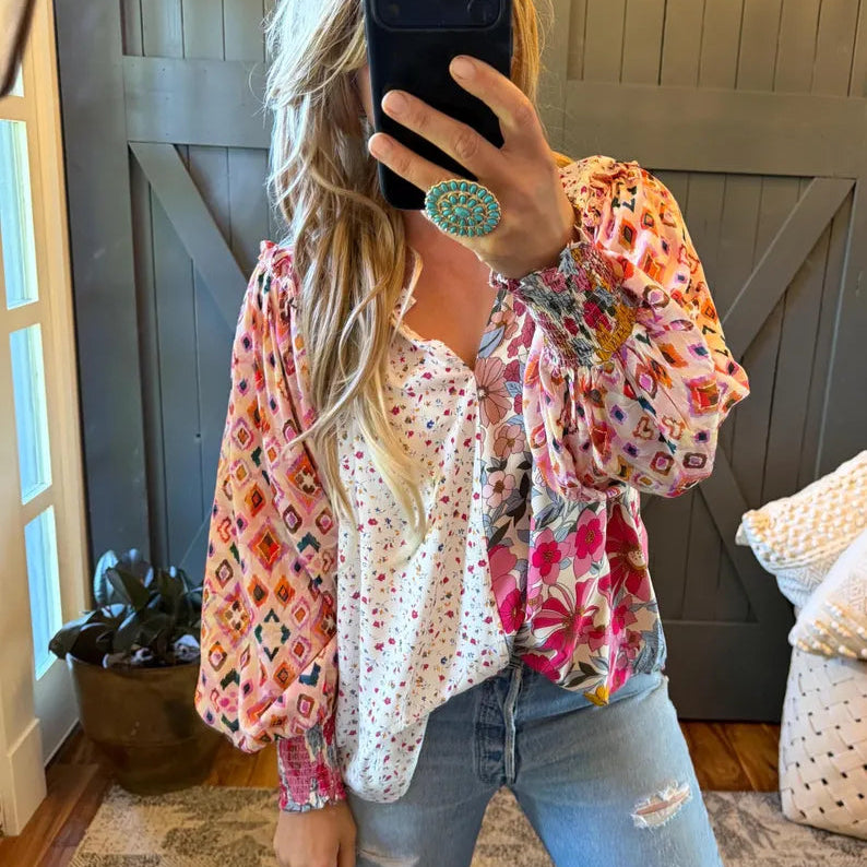 A woman takes a mirror selfie wearing a layered bohemian-style blouse with a white polka-dot inner top and a colorful patterned outer layer featuring pink, orange, and yellow floral and geometric motifs, paired with distressed light-wash jeans.