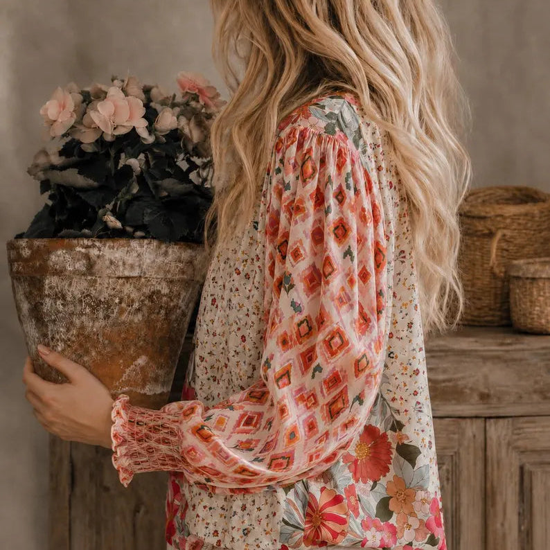 A woman shown from the back and side wearing a long-sleeve blouse with a cream base and colorful floral and geometric patterns, holding a rustic planter in a warm indoor setting.