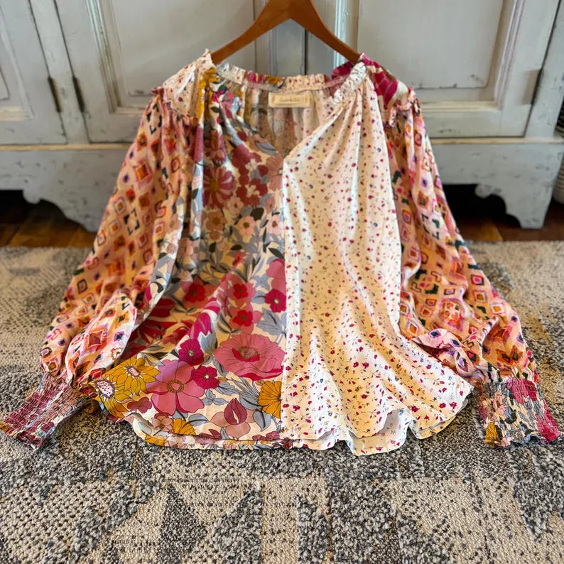 A long-sleeve blouse hanging on a wooden hanger, featuring a split design with a multicolor floral print on one side and a white base with small red and yellow polka dots on the other. The blouse is displayed on a patterned rug in front of a white cabinet.
