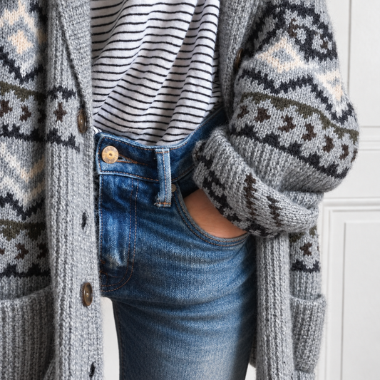 A close-up of a person wearing a gray knit cardigan with a dark and cream geometric pattern, a black and white striped t-shirt, and blue jeans.