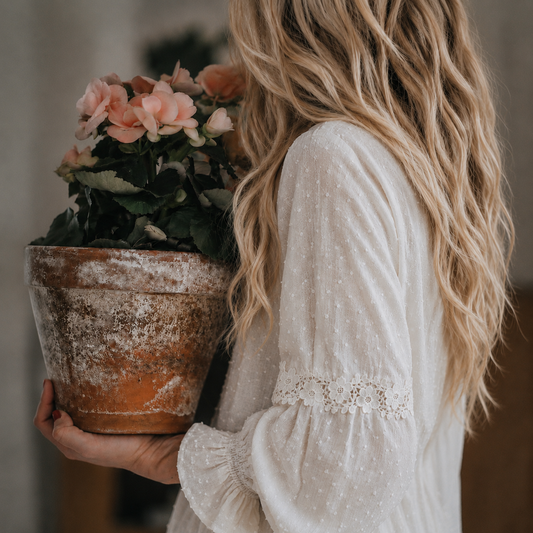 A woman with long wavy blonde hair is shown from behind, holding a rustic terracotta pot with pink flowers. She is wearing a white long-sleeve blouse with subtle polka dots and lace trim on the cuffs.