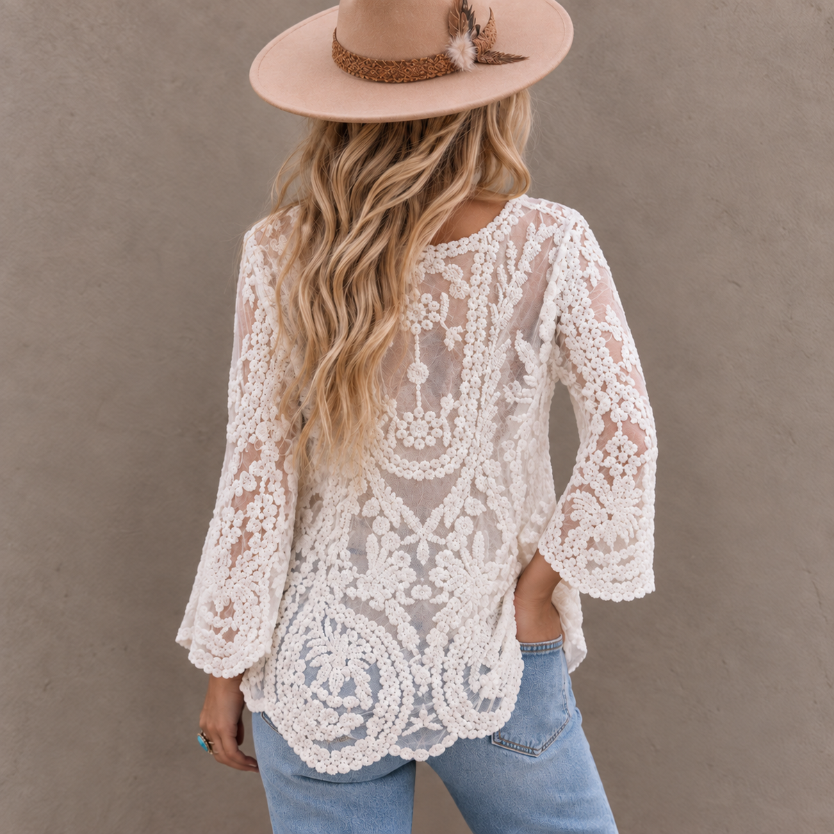 A woman shown from the back wearing a white lace long-sleeve top with a scalloped hem, paired with light blue jeans and a tan wide-brim hat, standing against a textured gray wall.