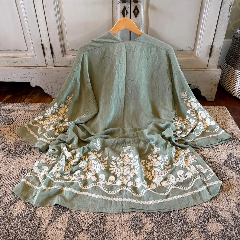 A sage green linen kimono-style cover-up with white floral embroidery and lace trim, displayed on a wooden hanger on a patterned rug in front of a distressed white cabinet.