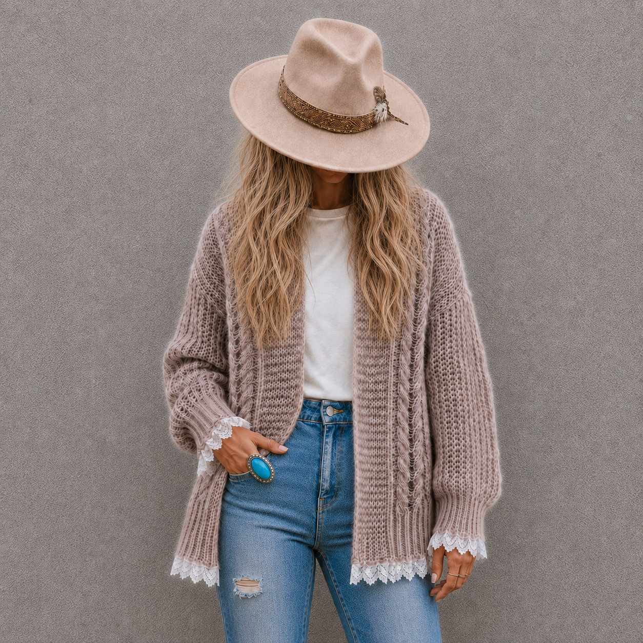 A woman wearing a chunky knit cardigan, a beige wide-brim hat, a white t-shirt, and blue jeans, standing against a textured gray wall.