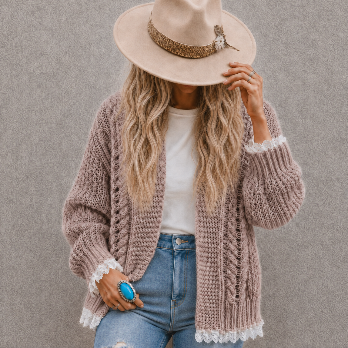 A woman wearing a chunky knit cardigan in a dusty mauve-pink color with white lace trim, a beige wide-brim hat, a white t-shirt, and blue jeans, standing against a textured gray wall.
