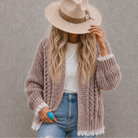A woman wearing a chunky knit cardigan in a dusty mauve-pink color with white lace trim, a beige wide-brim hat, a white t-shirt, and blue jeans, standing against a textured gray wall.