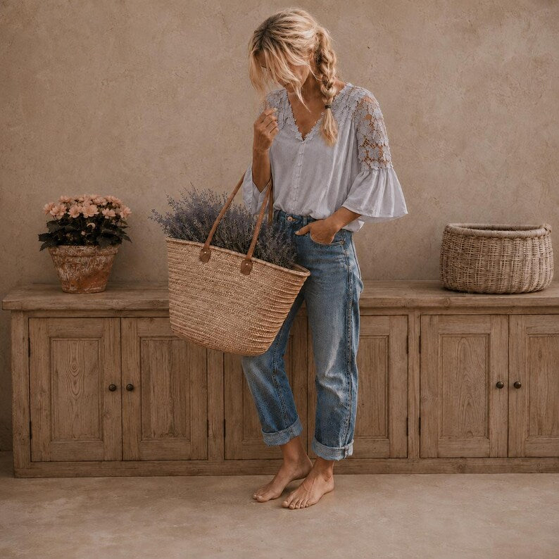 A woman stands barefoot in a rustic room, holding a large woven straw tote basket with lavender inside. She wears a light blue lace-sleeve blouse and blue jeans.