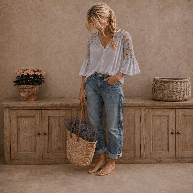 A woman stands barefoot in front of a rustic wooden cabinet, wearing a white lace-sleeve blouse and blue jeans, while holding a woven basket filled with lavender. The setting has a warm, earthy tone with a textured wall and a potted plant on the cabinet.