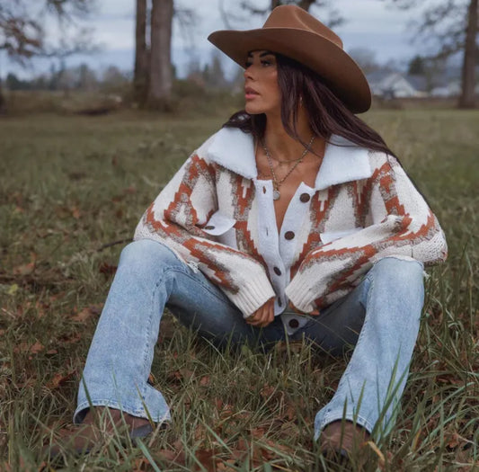 Woman wearing a patterned cardigan, jeans, and a brown hat sitting in a grassy field.