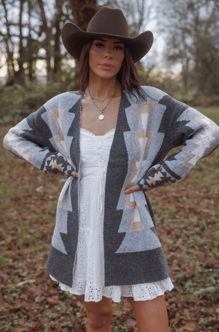 Woman wearing a patterned cardigan over a white dress with a cowboy hat outdoors.