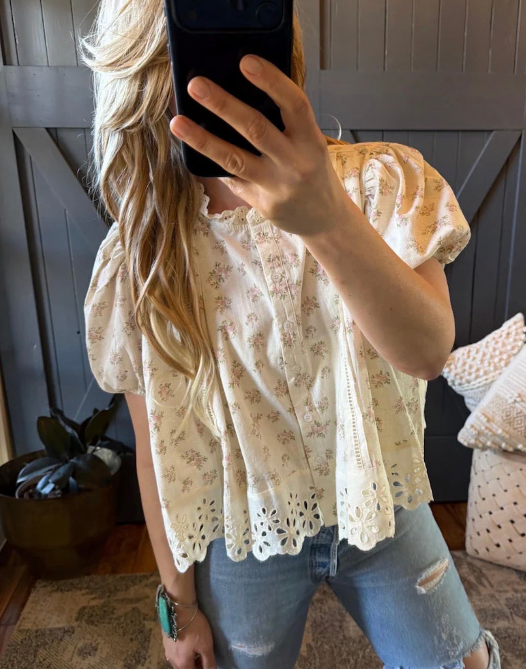 Person wearing a floral blouse and jeans, taking a mirror selfie.
