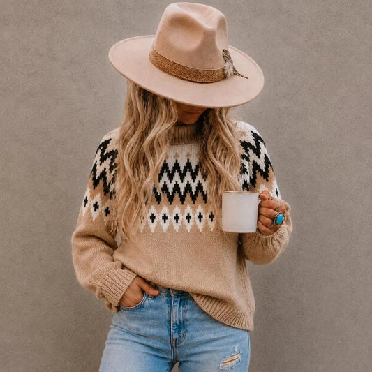 A woman wearing a beige knit sweater with a black and white geometric pattern, a beige wide-brim hat, and light blue jeans, holding a white mug against a textured gray wall.