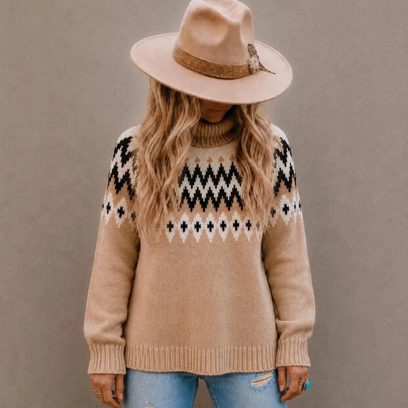 A woman wearing a beige knit turtleneck sweater with a black and white geometric pattern across the chest, styled with a matching beige wide-brim hat and light blue jeans, set against a plain taupe background.