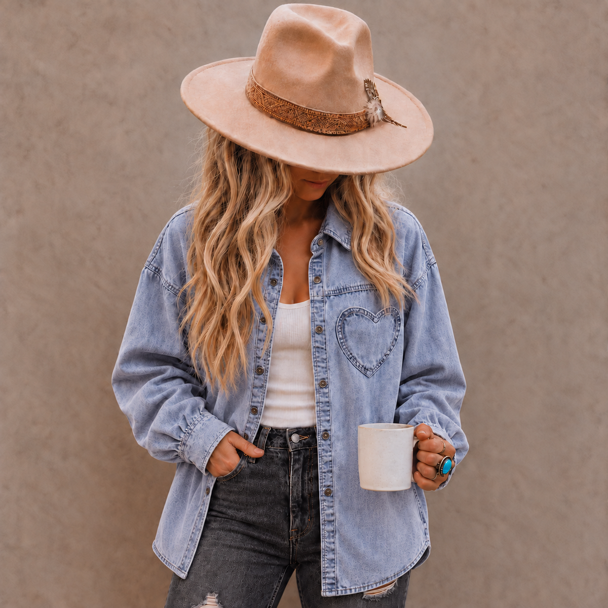 A woman wearing a light blue denim shirt with a heart patch on the pocket, a beige wide-brim hat, a white tank top, and dark gray jeans, holding a white mug against a textured brown wall.