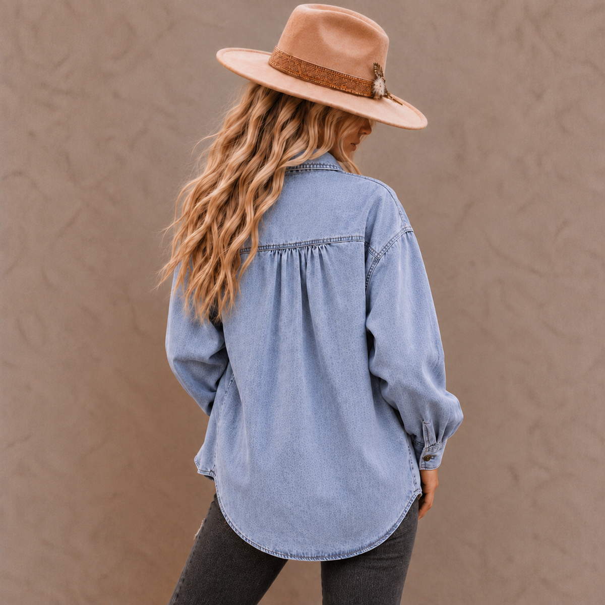 A woman shown from the back wearing a light blue denim shirt with long sleeves, gathered at the back, paired with dark gray pants and a tan wide-brim hat against a textured beige wall.