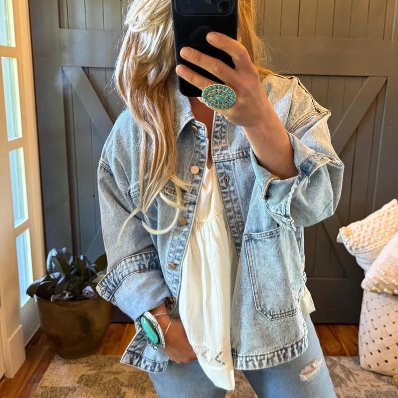 A woman takes a mirror selfie wearing a light-wash distressed denim jacket over a white top and matching light-wash jeans. She is wearing a large turquoise ring and a turquoise watch, with a dark wood door and indoor room visible in the background.