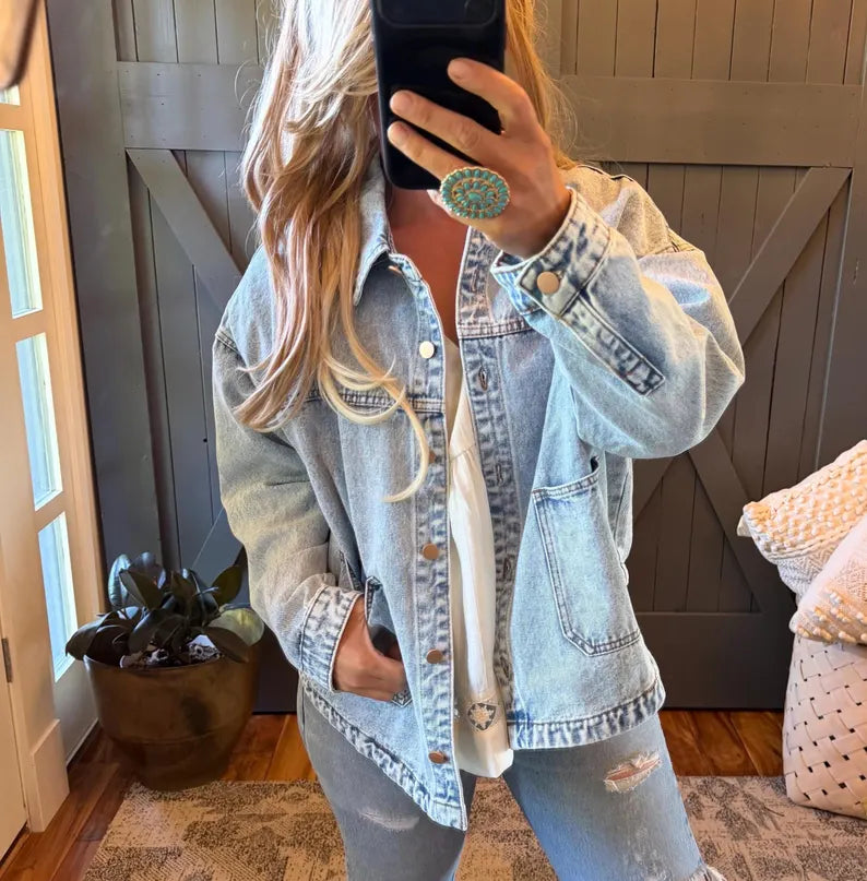 A woman takes a mirror selfie wearing an oversized light-wash blue denim jacket over a white top and matching distressed jeans. She is standing in front of a dark wood barn-style door with a plant and pillows visible in the room.