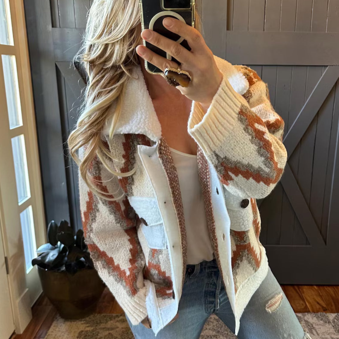 Person wearing a patterned cardigan taking a mirror selfie indoors.