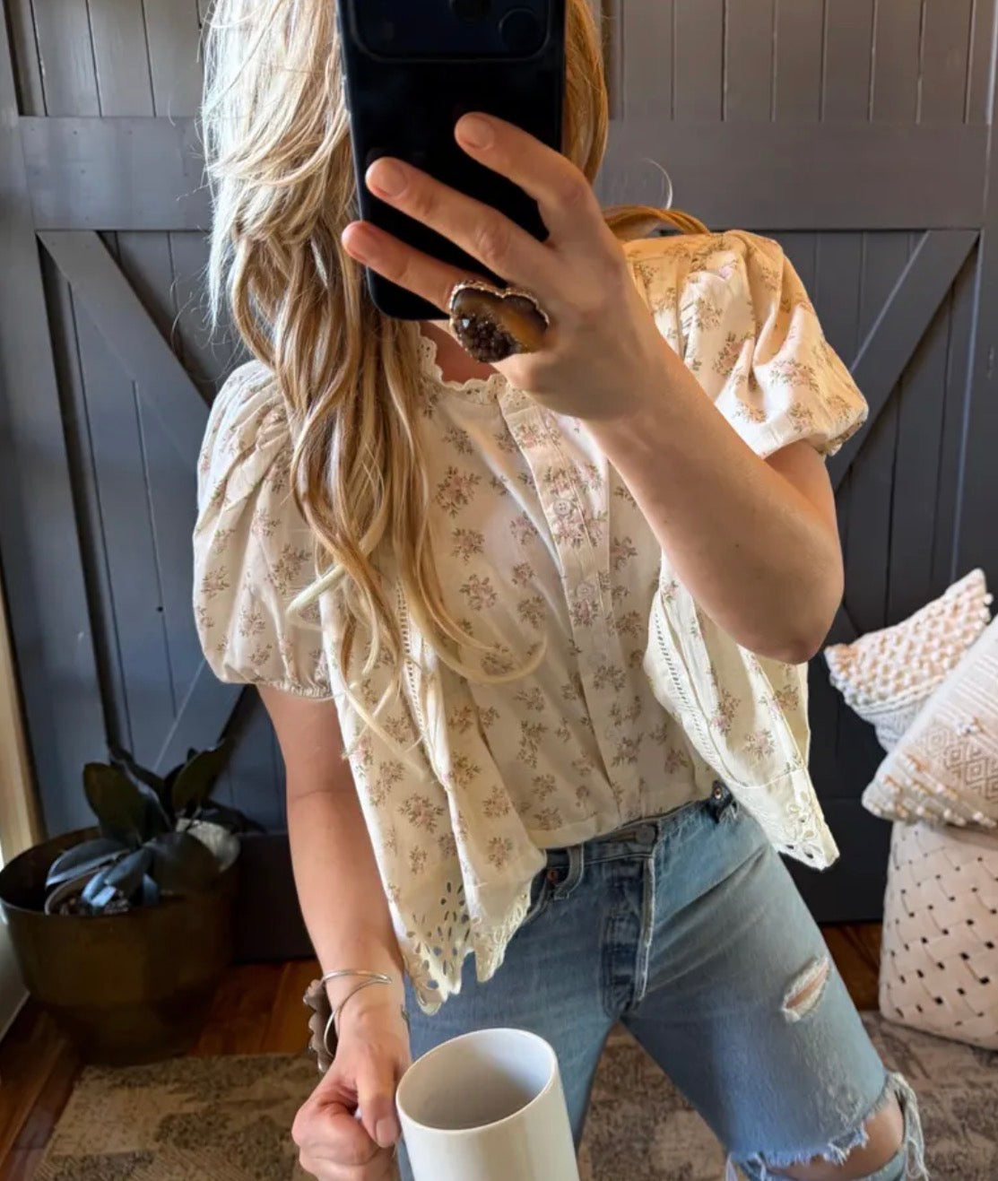 Person taking a mirror selfie wearing a floral blouse and jeans, holding a phone and a cup.