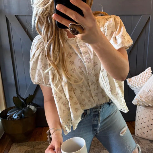 Person taking a mirror selfie wearing a floral blouse and jeans, holding a phone and a cup.