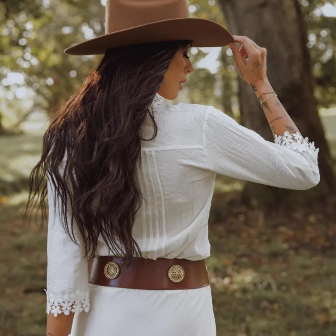 Woman wearing a white blouse, brown belt, and cowboy hat in a forest setting