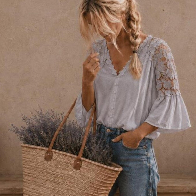 A woman wearing a light gray lace-sleeve blouse and blue jeans carries a large woven straw tote bag filled with lavender, standing against a textured beige wall.