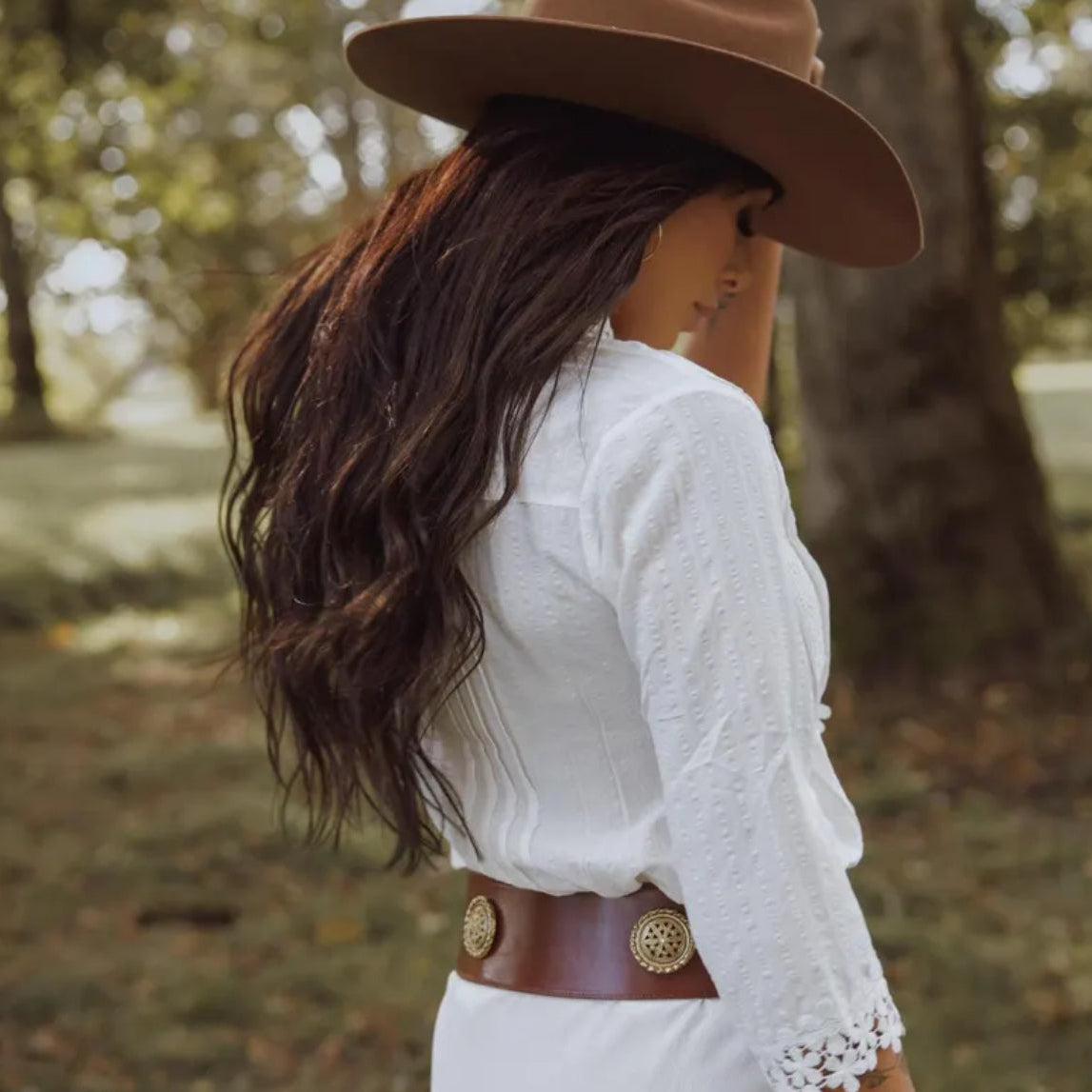 Woman wearing a white blouse, brown hat, and leather belt with a gold buckle in a natural setting.