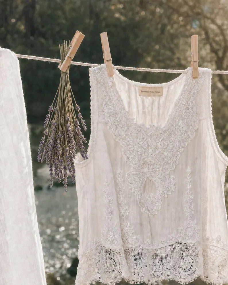 A white sleeveless lace blouse hanging on a clothesline outdoors, clipped with wooden clothespins next to a bunch of dried lavender. A matching white garment is partially visible on the left.