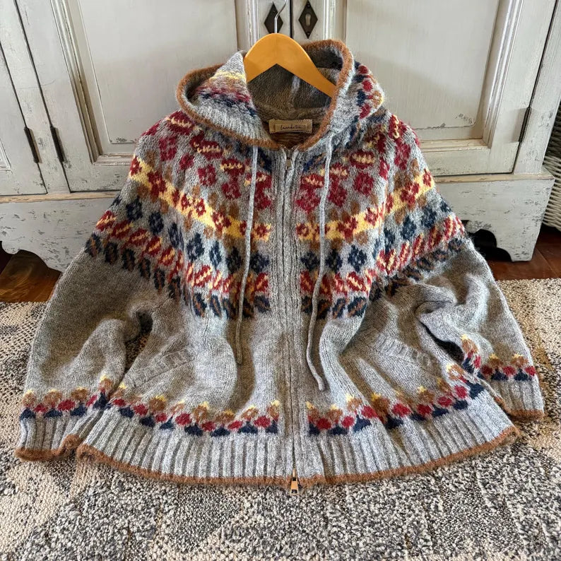 A knitted zip-front hooded sweater with a multicolor Fair Isle pattern on a heather gray base, hanging on a wooden hanger on a textured rug in front of a distressed white cabinet.